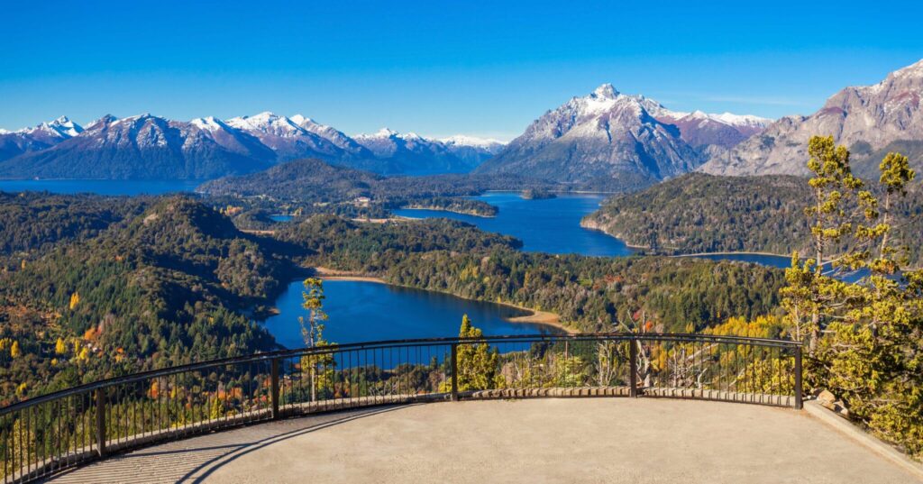 Mirador panorámico de la Cordillera de los Andes y lagos en Bariloche, con un bosque de otoño y un cielo despejado de fondo.