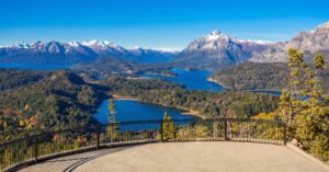 Mirador panorámico de la Cordillera de los Andes y lagos en Bariloche, con un bosque de otoño y un cielo despejado de fondo.