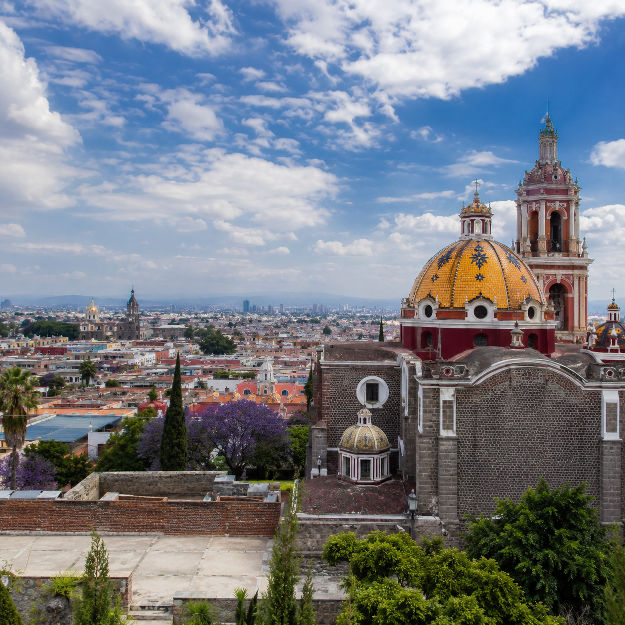 Vista panorámica desde lo alto mostrando las cúpulas decoradas y la torre del Santuario de la Virgen de los Remedios en Cholula, con la ciudad de Puebla extendiéndose al fondo bajo un cielo parcialmente nublado.