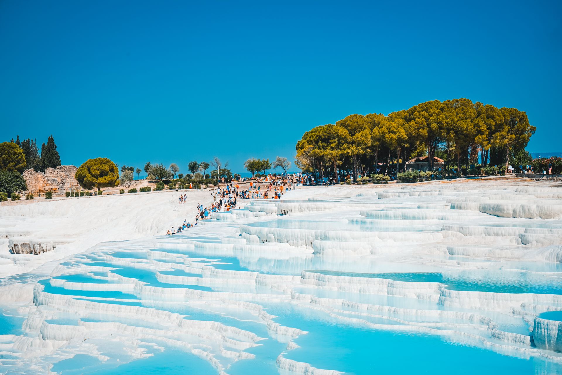 Terrazas de travertino blanco con piscinas de agua turquesa en Pamukkale, Turquía, con turistas paseando y árboles al fondo.