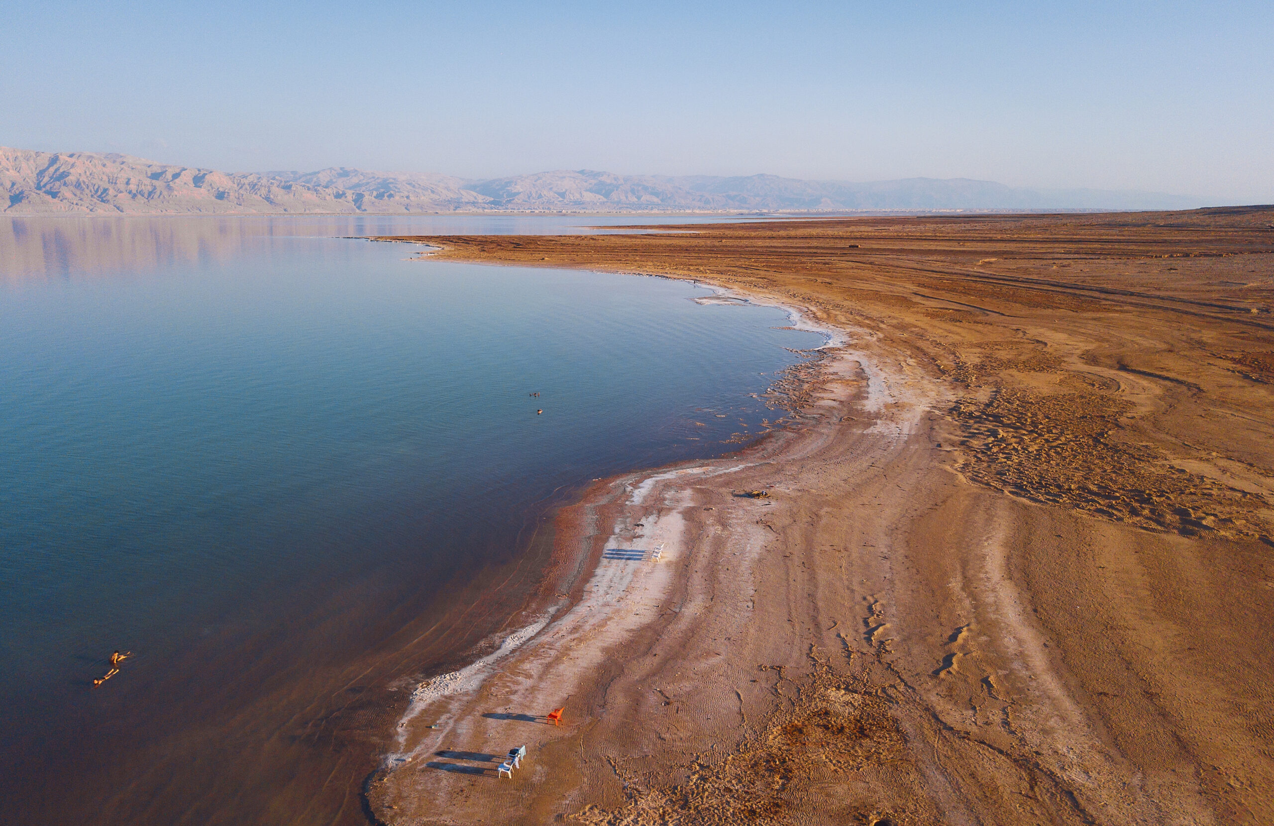 Vista aérea panorámica de las aguas tranquilas del Mar Muerto en Israel con depósitos de sal en la orilla y montañas desérticas al fondo.