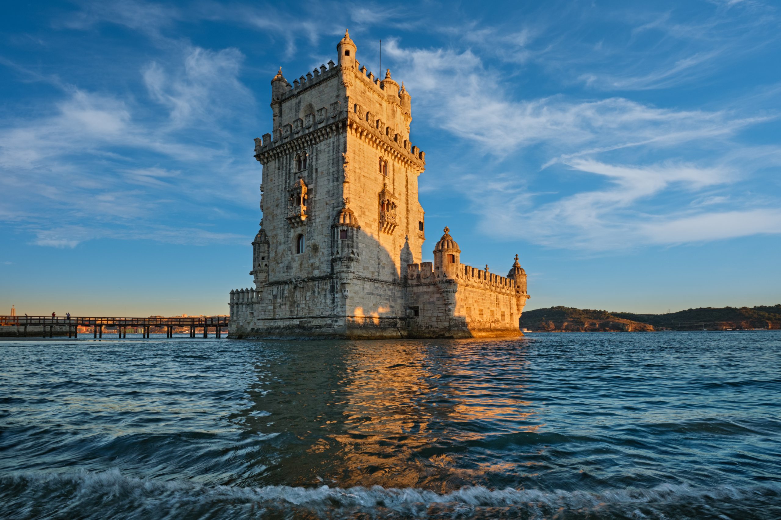 Vista de la icónica Torre de Belém a orillas del río Tajo en Lisboa durante el atardecer, con el agua en primer plano y un cielo azul con nubes suaves.