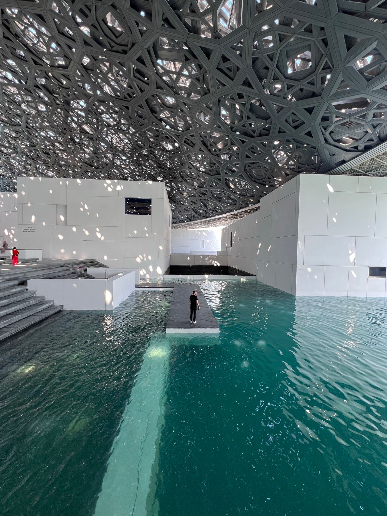 Persona de pie sobre pasarela flotante en las aguas del Museo Louvre Abu Dhabi, bajo la icónica cúpula de malla geométrica[
